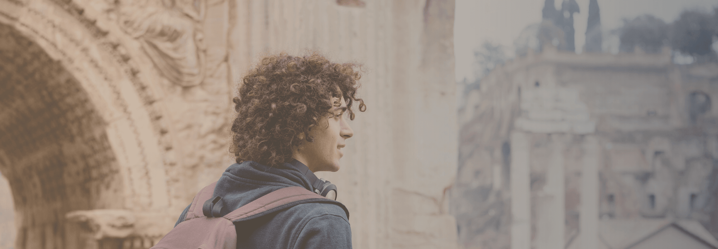 Young man with curly, brown hair wearing backpack while walking in front of Roman ruins